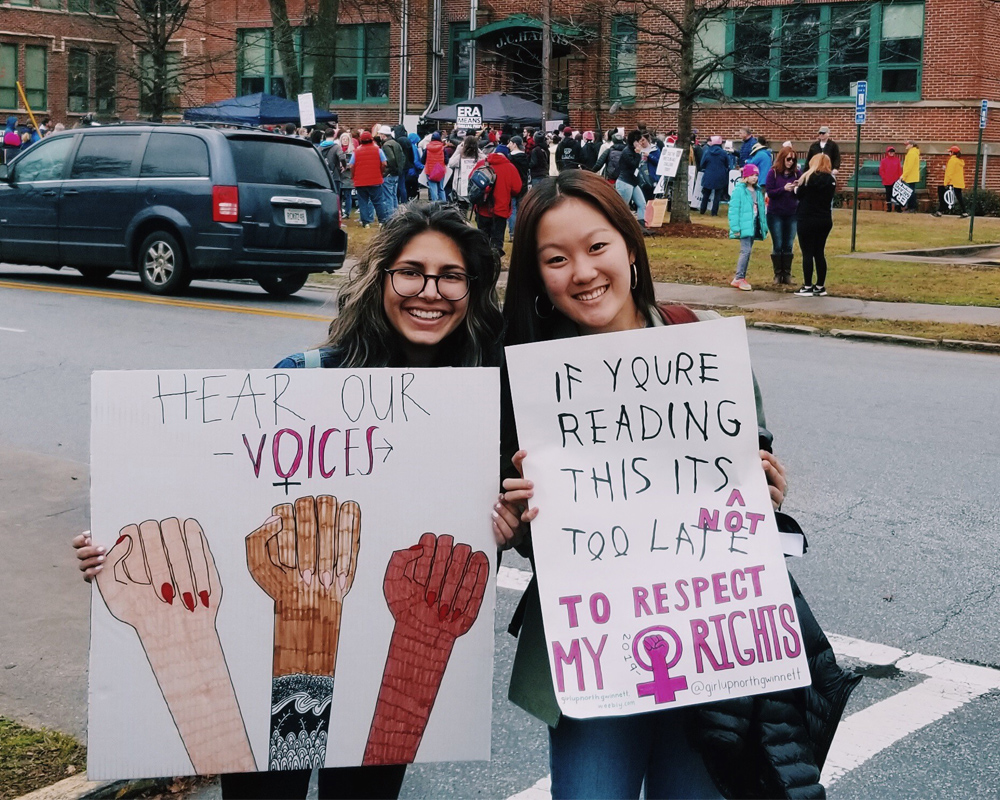 two girls are in the women march, tey are smile and holding sign "hear our voice" and "if you re reading this its not too late"