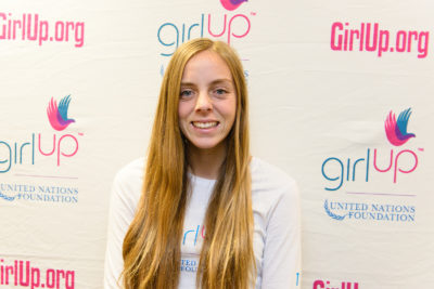 Alexis Kallen_2013-2014 Teen Advisor (close angle headshot, a picture little blurry ) a teen girl wearing her girl up white shirt with her smiley face facing the camera, and background is girlup.org board
