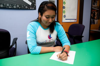 Angie Partida_The founding class of Teen Advisors(straight angle, but not a clear picture ) a teen girl wearing her girl up blue long shirt with her smile face facing down and holding a pen on her notebook, and background is an office