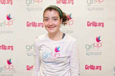 Delia Friel_2012-2013 Class Teen Advisors (close angle headshot, a picture little blurry ) a teen girl wearing her girl up white shirt with her smiley face facing the camera, and background is girlup.org board