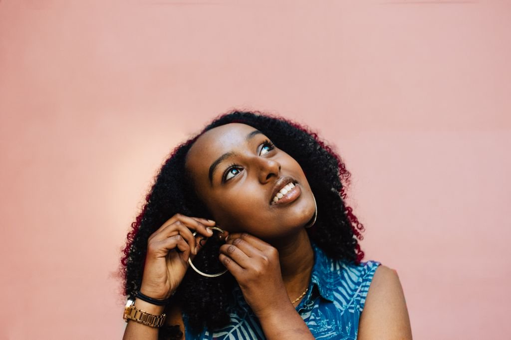 a girl is fixing her earring on her ear and looking up to the sky with a smile and the background is pink