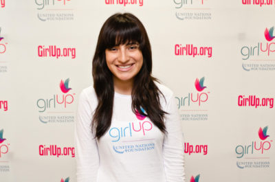Ines Renique Hometown: New York, NY_2012-2013 Class Teen Advisors (close angle headshot, a little blurry picture ) a teen girl wearing her girl up white shirt with her smiley face facing the camera, and background is girlup.org board