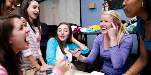 5 girls in the picture, girl in the center has her one finger pointing at her hand, and one finger on the other hand pointing at her girl cross her with big smile on her face, and everyone else are laughing