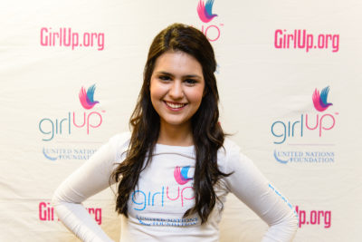 Julia Sumouske_2013-2014 Teen Advisor (close angle headshot, a picture little blurry ) a teen girl wearing her girl up white shirt with her smiley face facing the camera, and background is girlup.org board