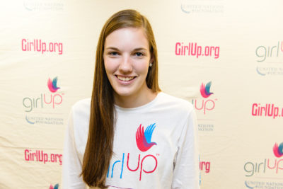 Kate McCollum_2013-2014 Teen Advisor (close angle headshot, a picture little blurry ) a teen girl wearing her girl up white shirt with her smiley face facing the camera, and background is girlup.org board