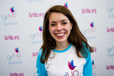 Katherine Cochran The founding class of Teen Advisors(close angle, but not clear picture headshot ) a teen girl wearing her girl up blue long shirt with her smiley face facing the camera, and background is girlup.org board