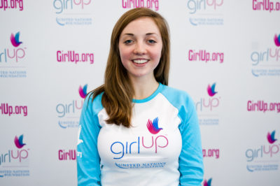 Lily Kaplan,The founding class of Teen Advisors(close angle, but not clear picture headshot ) a teen girl wearing her girl up blue long shirt with her smiley face facing the camera, and background is girlup.org board