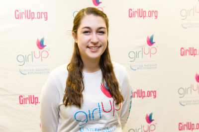 Lindsay Schrier_2013-2014 Teen Advisor (close angle headshot, a picture little blurry ) a teen girl wearing her girl up white shirt with her smiley face facing the camera, and background is girlup.org board