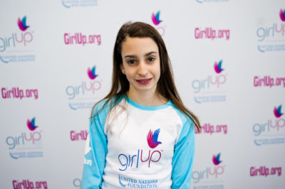 Olivia Layton,The founding class of Teen Advisors(close angle, but not clear picture headshot ) a teen girl wearing her girl up blue long shirt with her smiley face facing the camera, and background is girlup.org board