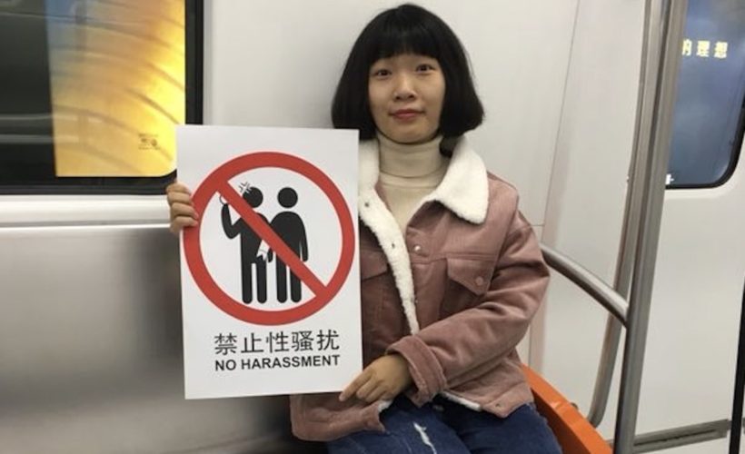 a Chinese girl is sitting down on the metro seat, facing the camera and holding a sign "no harassment."