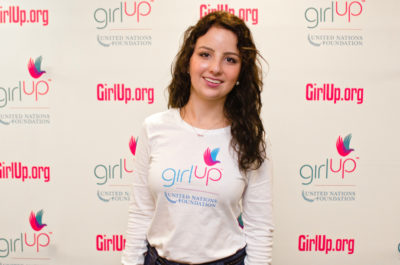 Sofia Stafford Hometown: New York, NY_2012-2013 Class Teen Advisors (close angle headshot, a little blurry picture ) a teen girl wearing her girl up white shirt with her smiley face facing the camera, and background is girlup.org board
