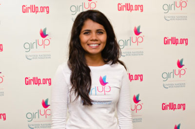 Sri Muppidi Hometown: Pleasanton, CA_2012-2013 Class Teen Advisors (close angle headshot, a little blurry picture ) a teen girl wearing her girl up white shirt with her smiley face facing the camera, and background is girlup.org board
