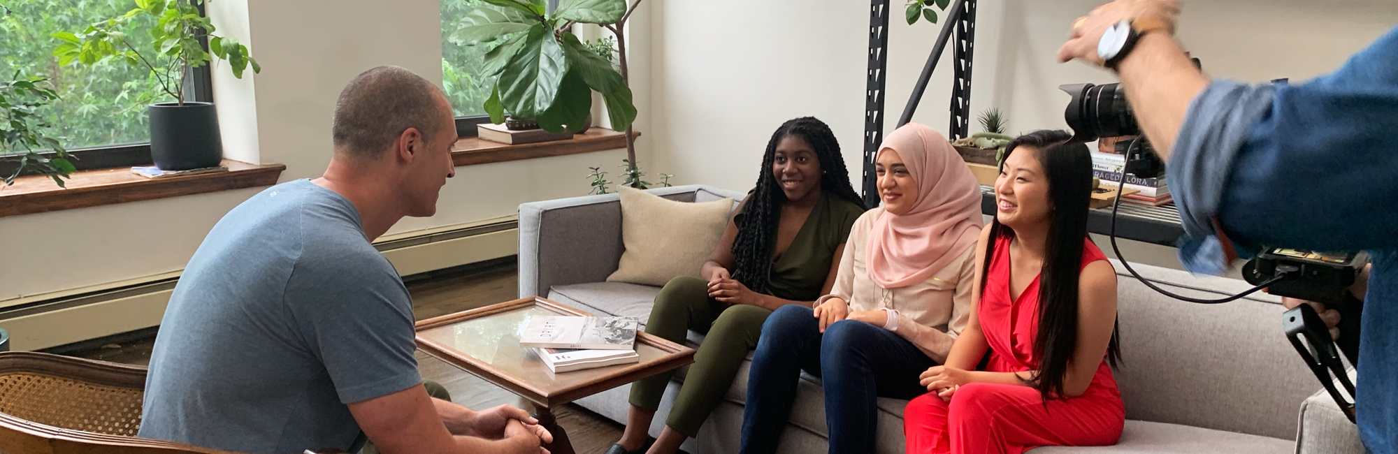 3 girl up girls on the couch and interviewing a man across them, and there is a camera on the right-hand side to recording them