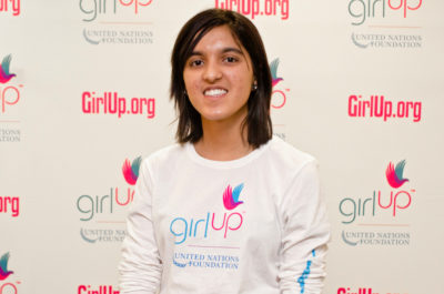 Tanya Devani, Hometown: Birmingham, AL_2012-2013 Class Teen Advisors (close angle headshot, a little blurry picture ) a teen girl wearing her girl up white shirt with her smiley face facing the camera, and background is girlup.org board