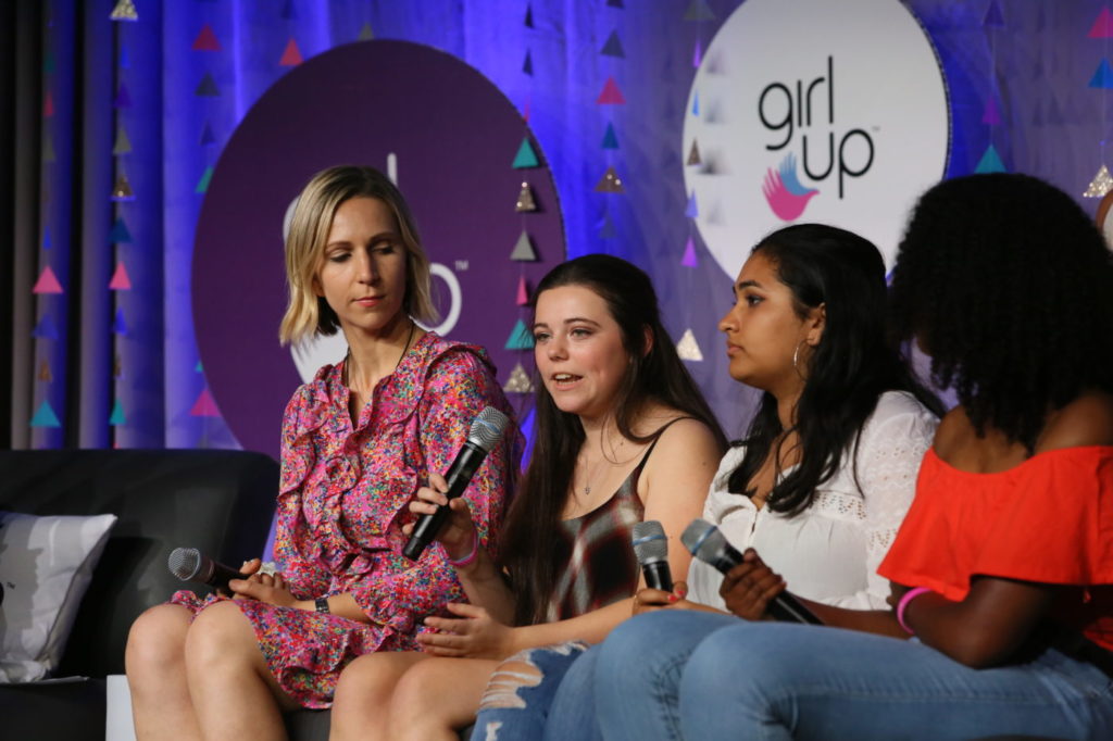 3 girls are sitting down on the couch on the stage and speaking with their mic on their hand