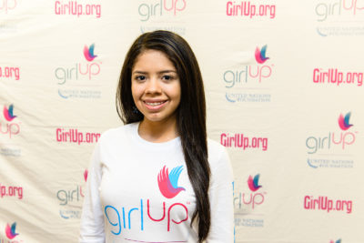 Valeria Hansen 2013-2014 Teen Advisor (close angle headshot, a picture little blurry ) a teen girl wearing her girl up white shirt with her smiley face facing the camera, and background is girlup.org board