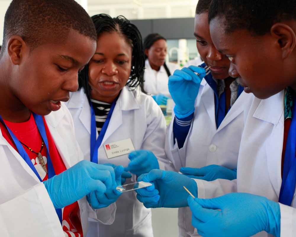 1 boys and 2 girl circle around with their white coat and looking the lap sample and listening to the teacher to explain