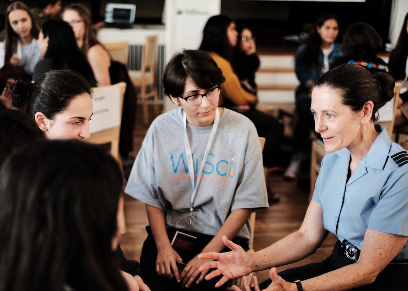 a woman is talking with 3 girl up girls in the pictures and the girls are wearing WiSci t-shirt in the centre is Becca Bean Teen Advisor Alumna and playing attention to the conversation