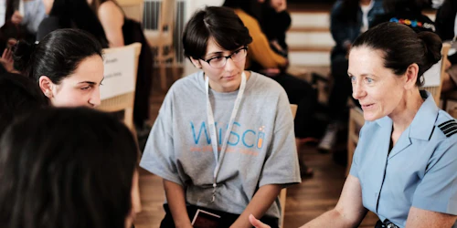 a woman is talking with 3 girl up girls in the pictures and the girls are wearing WiSci t-shirt in the centre is Becca Bean Teen Advisor Alumna and playing attention to the conversation