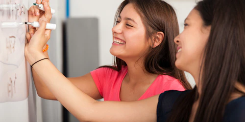 two girls are writing on the white board and laughing