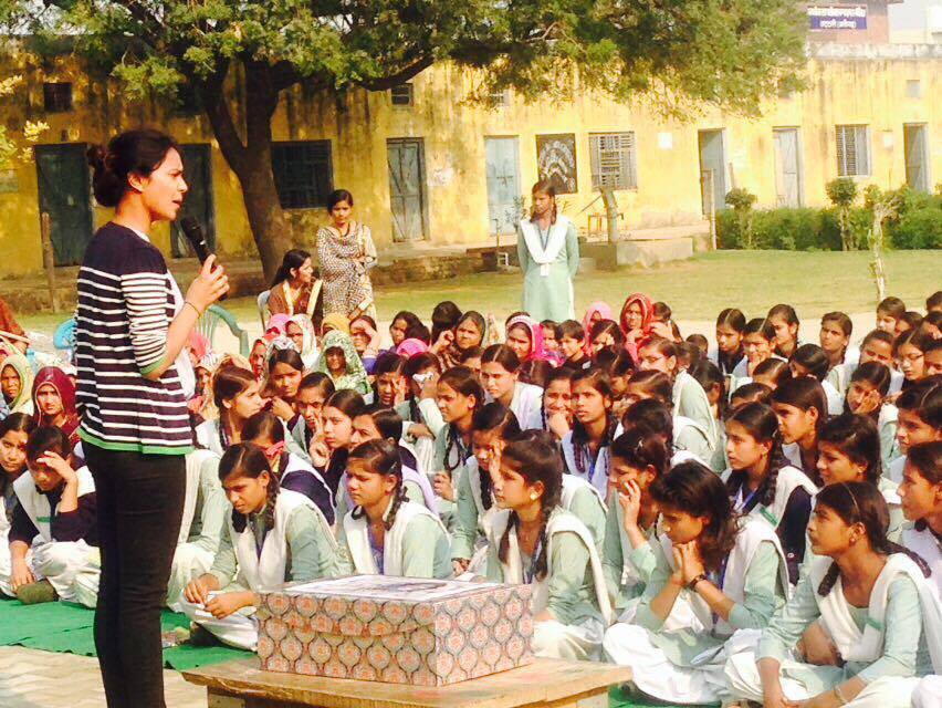 a woman standing and speaking in front of a group of girl students who are sitting on the group listen to her at the school