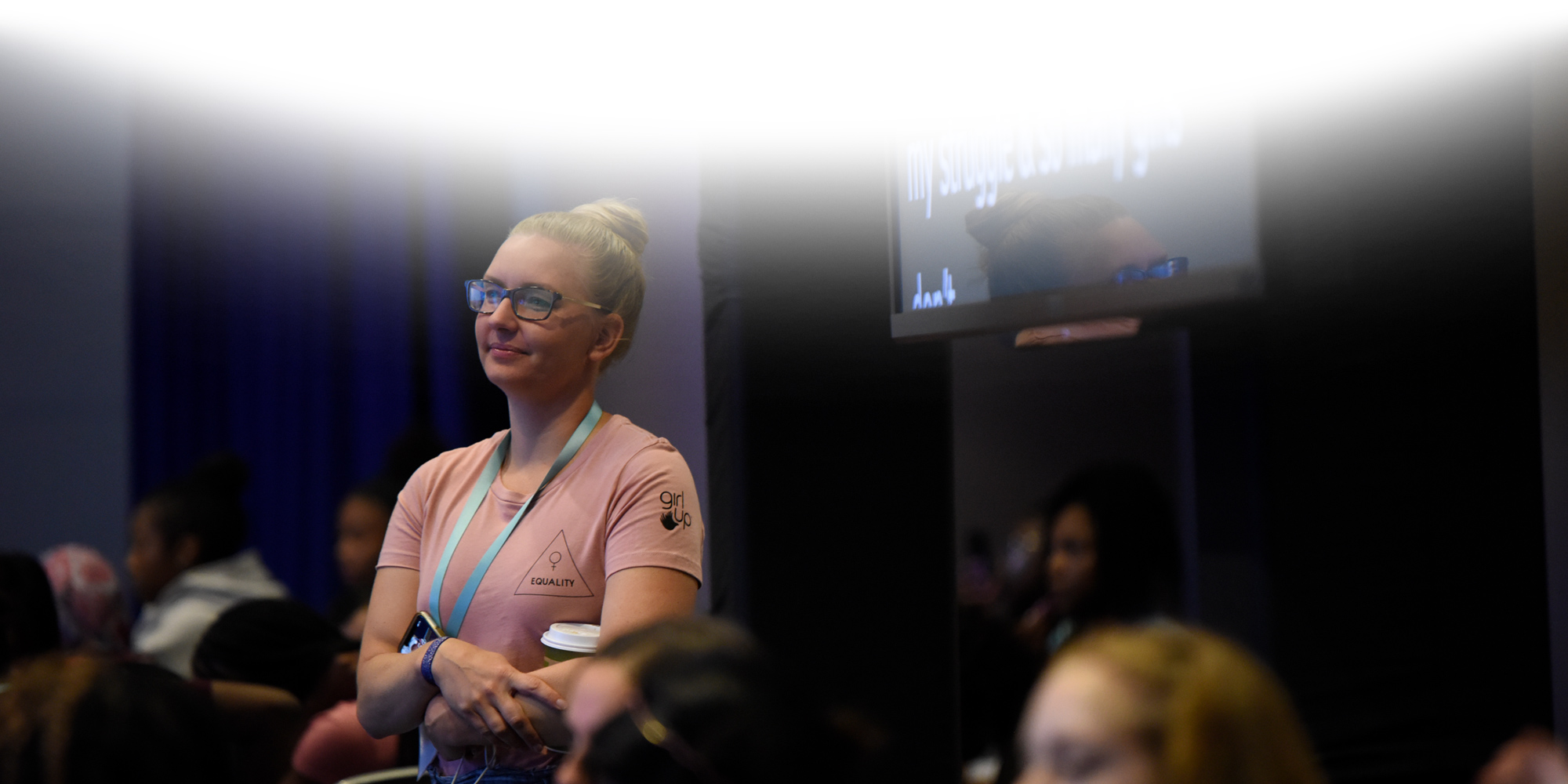a girl standing up among the crowd and smiling listening to the speaker at an event