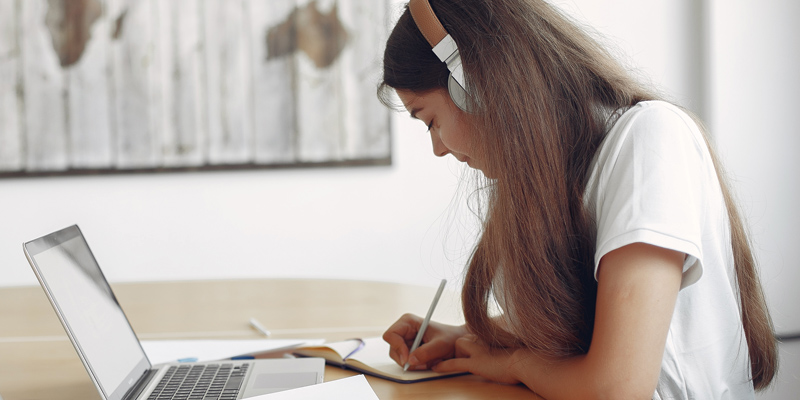 a girl writing notes with her headphone on and a laptop in front of her