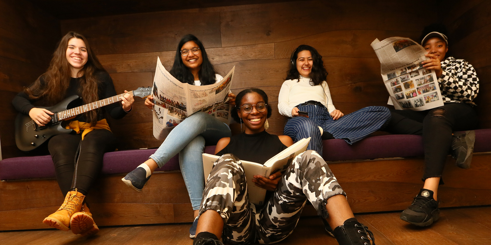 5 different ethnic girls in the picture holding books (sitting on the front floor) and holding guitar and newspaper (sitting on the back couch)
